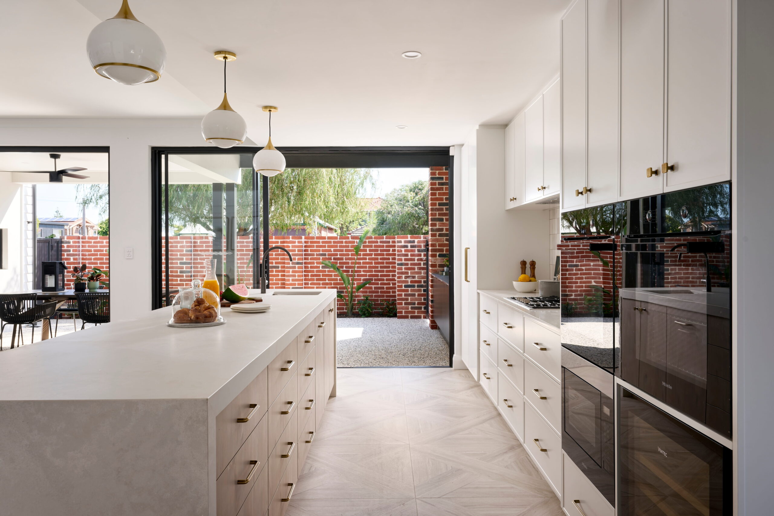 Custom kitchen in a Perth home with white cabinetry, brass hardware, stone island bench and globe pendants opening to a brick-walled alfresco. 