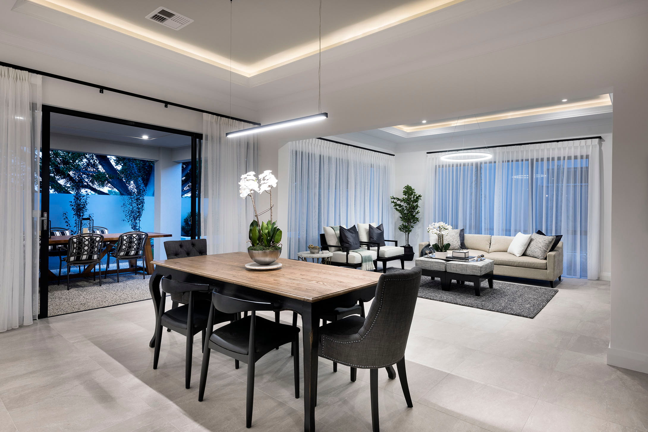 Spacious open-plan dining and living area in a luxury Perth home with cove ceiling lighting, large-format floor tiles and sheer floor-to-ceiling curtains.