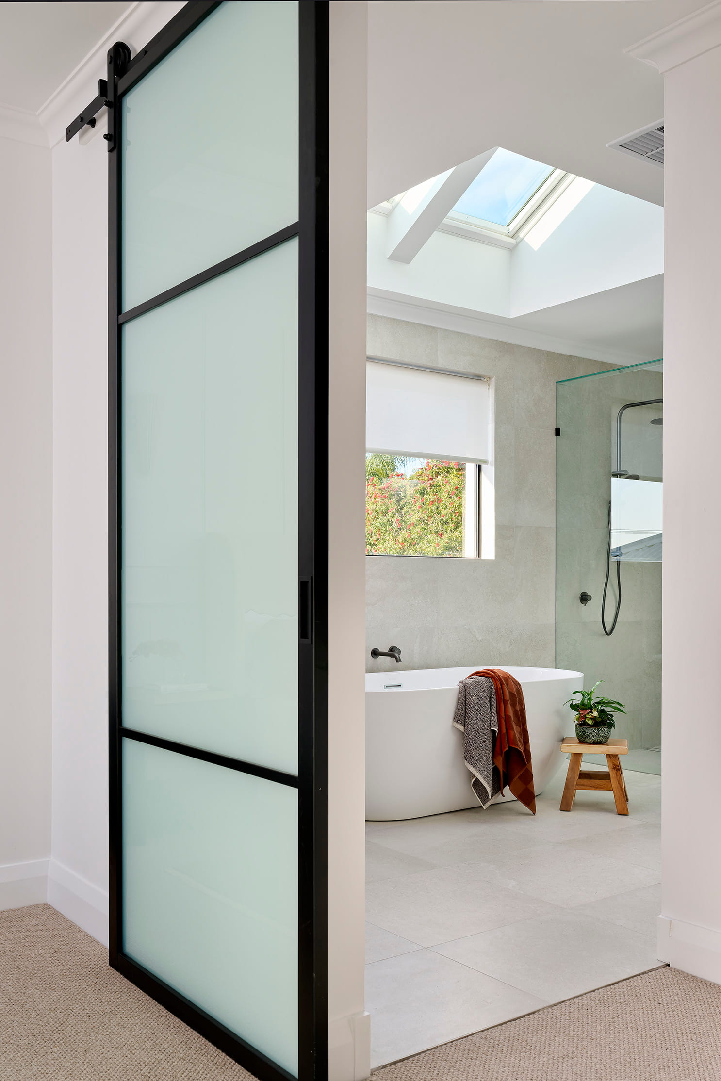 Ensuite bathroom in a custom Perth home with black-framed glass barn door, skylight, freestanding bath and stone wall tiles.